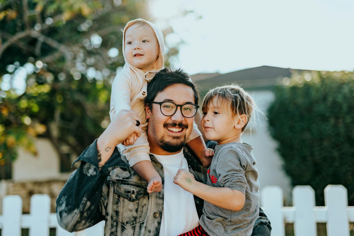 A smiling father holding two young children outside their home